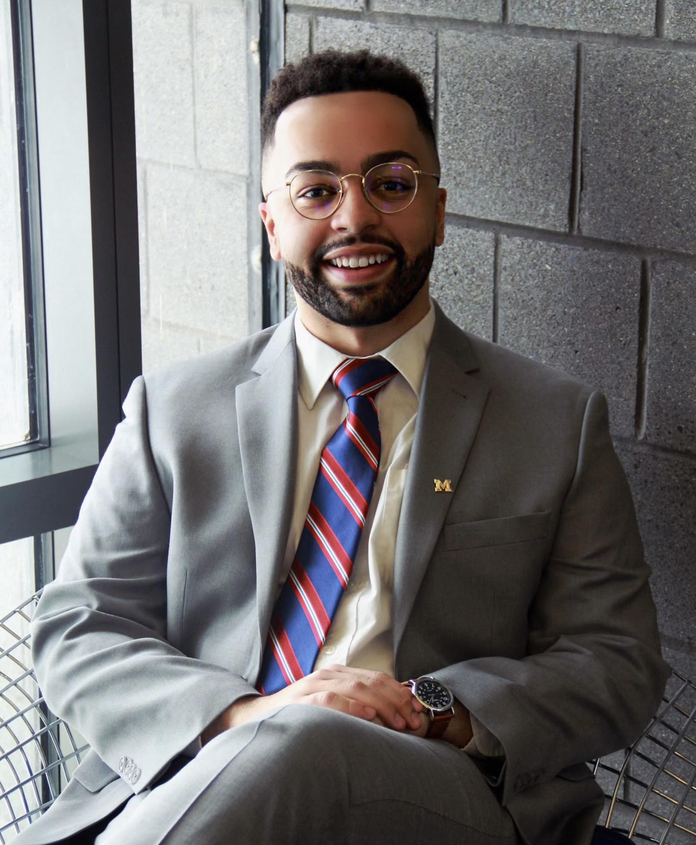 Person seated indoors in front of a modern, textured concrete wall. They are wearing a light gray suit, a white shirt, and a blue, red, and white striped tie. A round pin with the letter "M" is attached to their lapel. The person has short hair and wears round glasses.