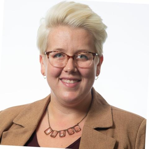 White woman with short blonde hair and glasses wearing a suit jacket and smiling on a white background