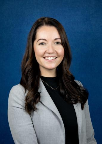 Portrait of a smiling person with long dark hair, wearing a gray blazer and black top, in front of a blue background.